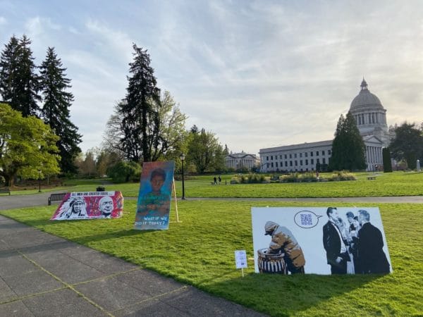Close up of three pieces of art next to sidewalk with grass, trees, and capitol building in background.